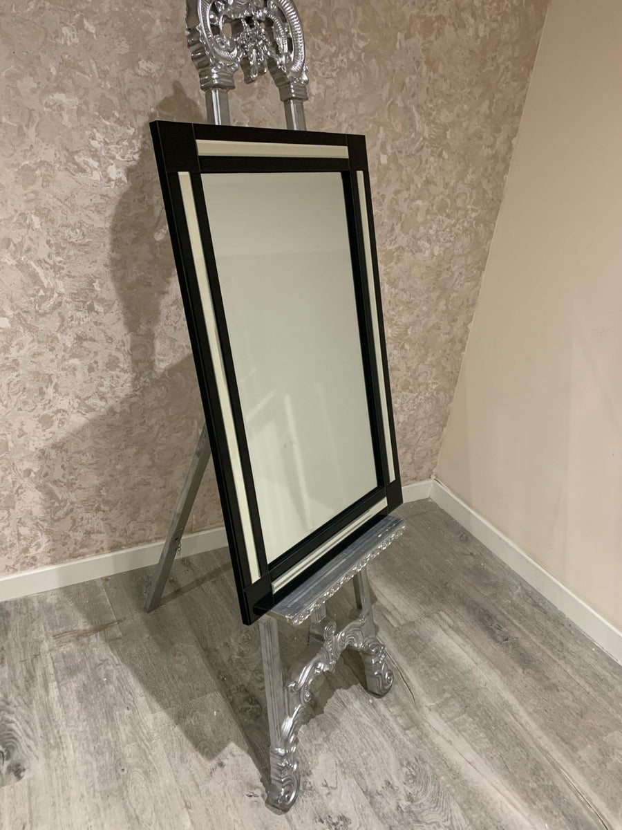Decorative mirror with a black frame and silver stand against a patterned wall.