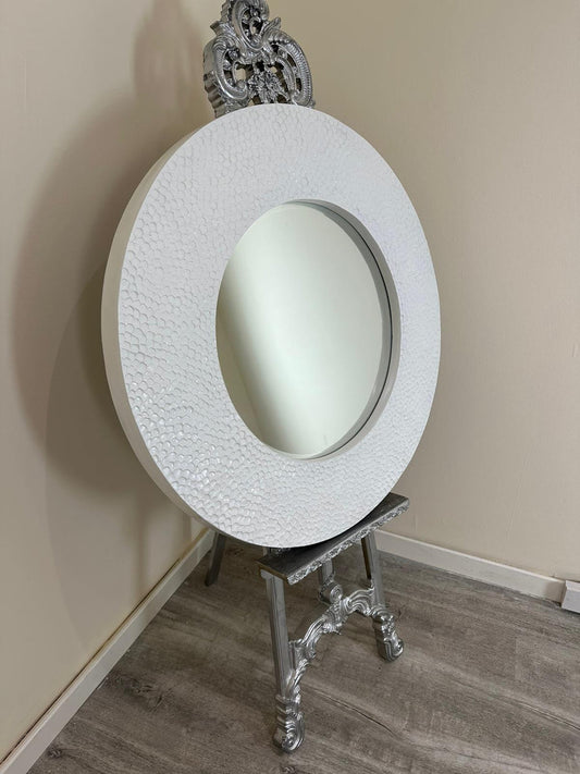 Decorative round mirror with intricate silver frame on a stand against a beige wall.