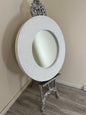 Decorative round mirror with intricate silver frame on a stand against a beige wall.